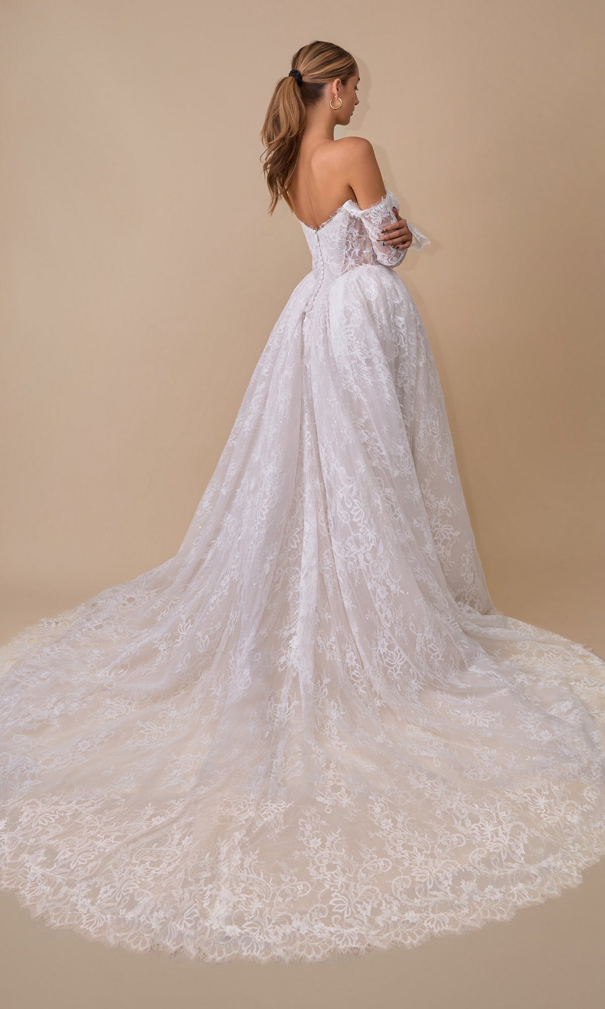 A woman stands facing away against a beige background, wearing the Andrea & Leo WL079 Long Wedding Dress—a white off-the-shoulder bridal gown featuring a long, flowing train and delicate lace detailing.